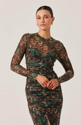 A woman is wearing a fitted, long-sleeved dress with a floral pattern in shades of green, brown, and beige. The dress features a sheer, mesh-like fabric at the upper chest and sleeves, with a ruched detail along the front and a center slit at the bottom.
